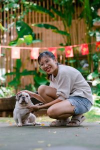 Smiling woman crouching next to a bulldog in a lush garden setting.