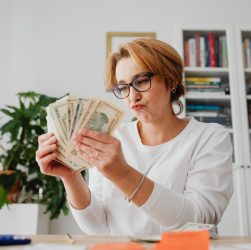 Woman wearing glasses, counting banknotes with a focused expression in a home office setting.