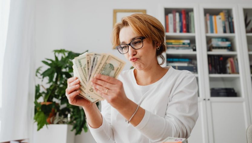 Easy Ways to Earn Extra Money and Ease Your Financial Stress 1 Woman wearing glasses, counting banknotes with a focused expression in a home office setting.