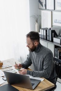 Easy Ways to Earn Extra Money and Ease Your Financial Stress 2 A bearded man counts dollar bills while seated at a desk with a laptop in an office setting.
