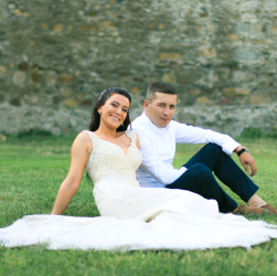 Full body of young bride and groom in elegant suit sitting together on green lawn in park and looking at camera