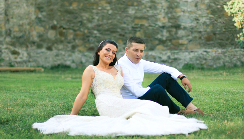 Full body of young bride and groom in elegant suit sitting together on green lawn in park and looking at camera